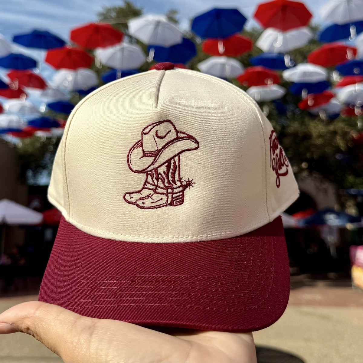 Cowgirl Boots Howdy Yall trucker hat in Natural / Maroon Bill with maroon cowboy boots and hat embroidery, held up under red, white and blue umbrellas