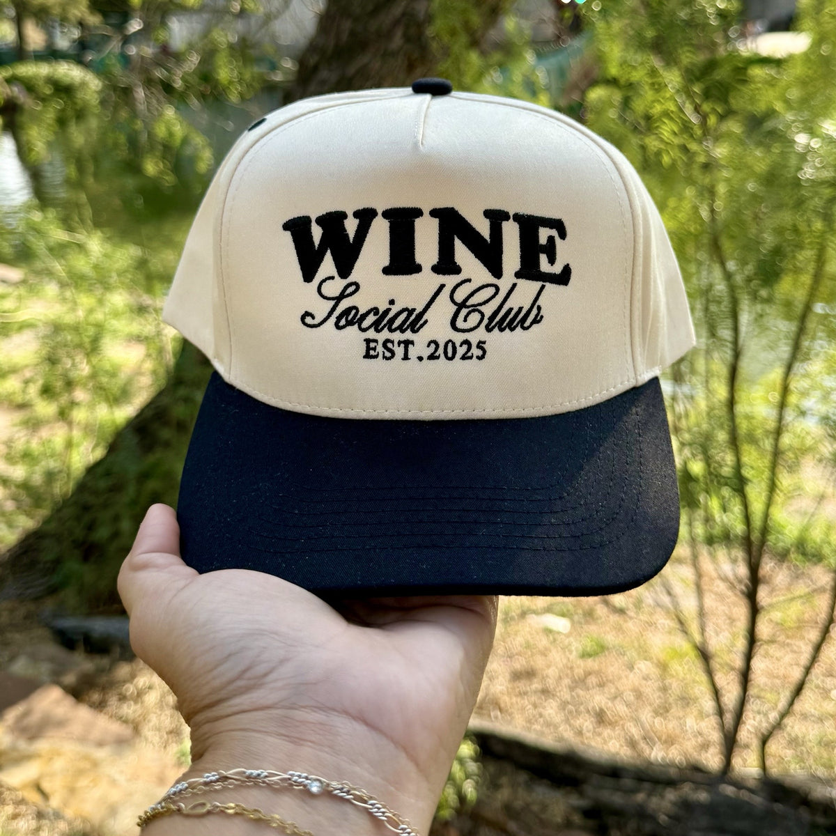 Wine Social Club trucker hat in Natural / Black Bill with black WINE Social Club EST.2025 lettering, held up outdoors