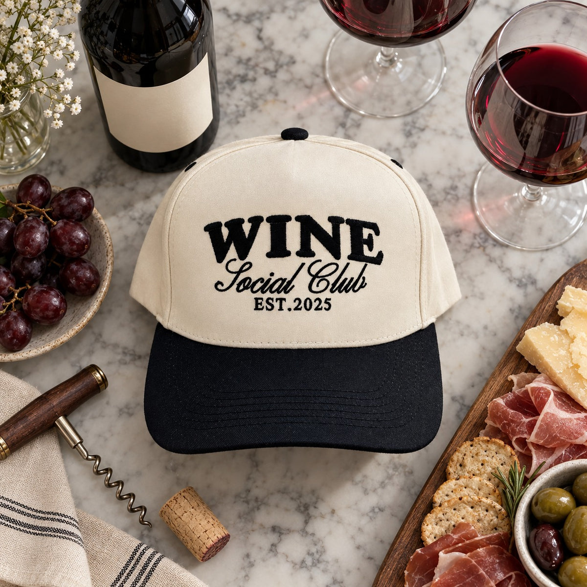 Wine Social Club trucker hat flat lay on marble with red wine, wine glasses, grapes, a corkscrew, and a charcuterie board