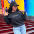 Woman wearing the black DTMF Puerto Rico Icons embroidered sweatshirt from DSY Lifestyle, standing on red steps with a colorful background.