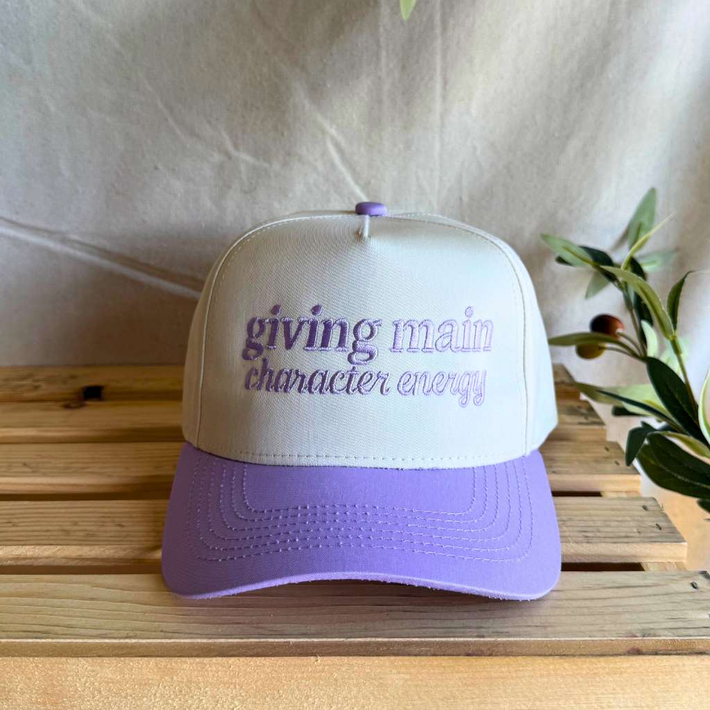 White and purple cap with text on a wooden surface Giving Main Character Energy Two Tone Hats - Inspired from the show Love Island