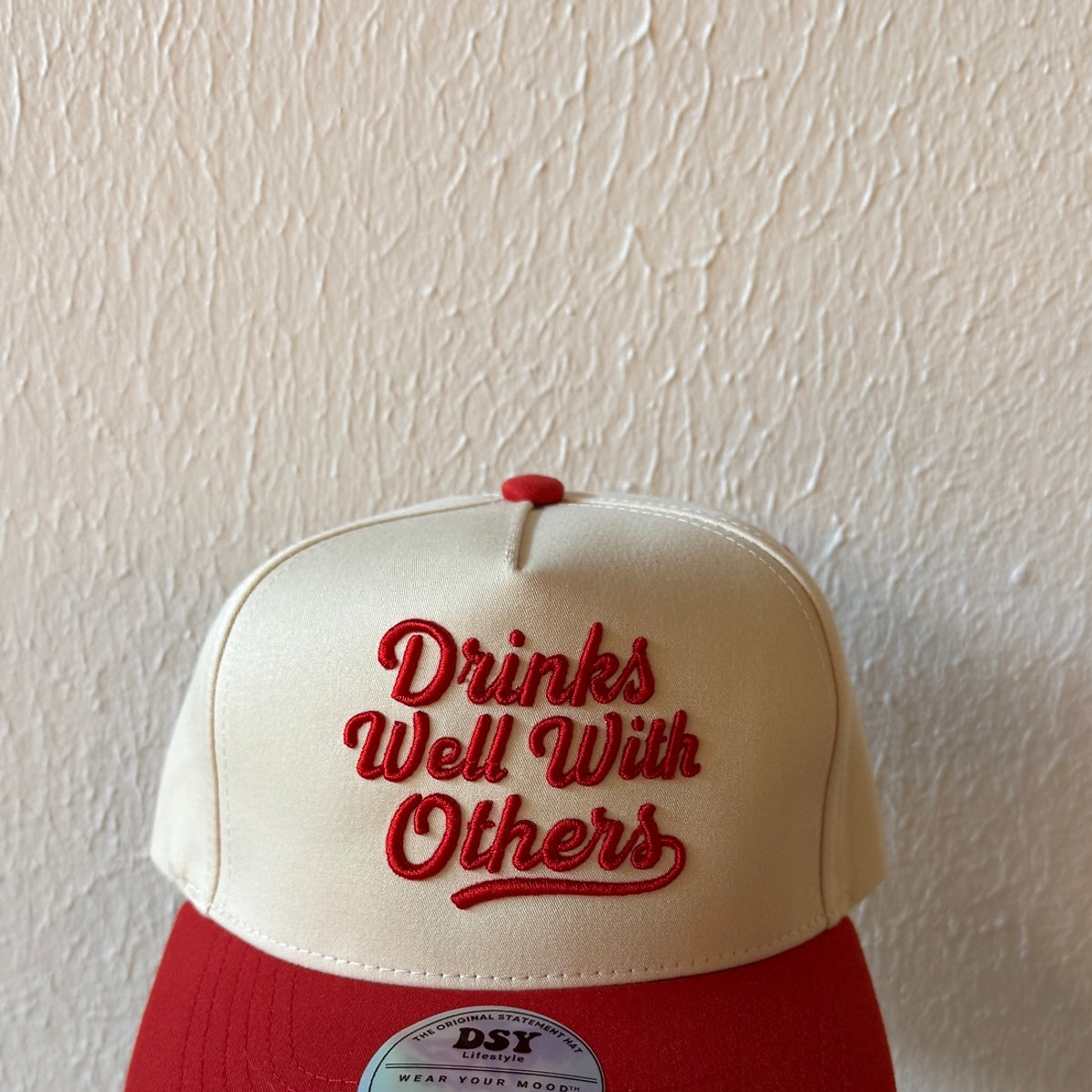 Close-up of Drinks Well With Others machine embroidery on a two-tone trucker hat