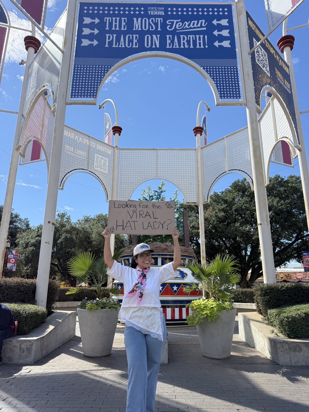 Daisy Delgado, the Texas Hat Lady, holding a Looking for the Viral Hat Lady sign under The Most Texan Place on Earth sign at the State Fair of Texas