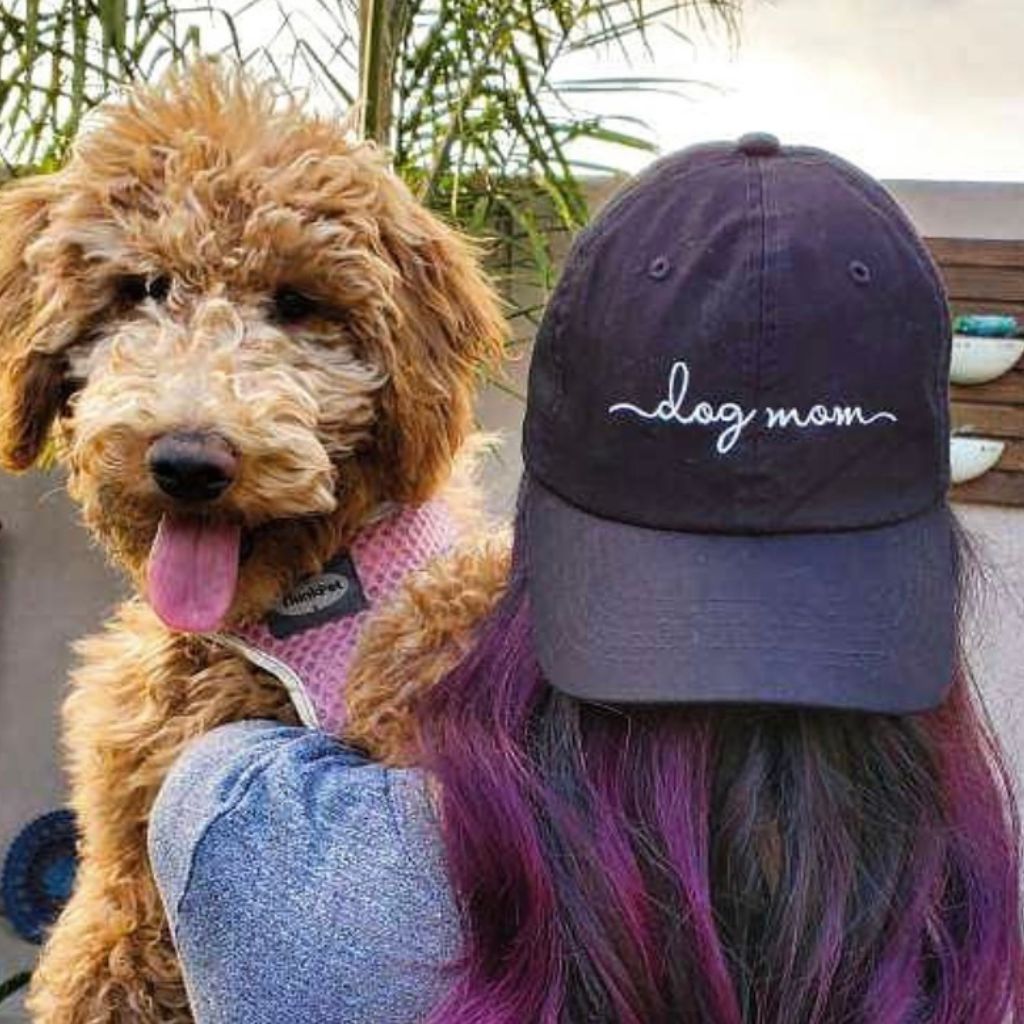 Female carrying a dog and wearing a black baseball cap embroidered with Dog Mom