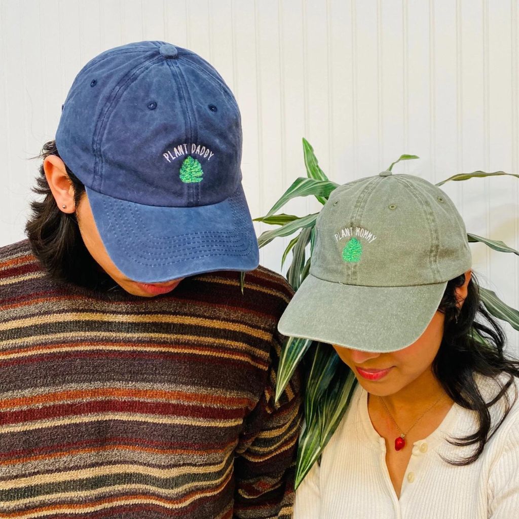 Male wearing a navy washed hat embroidered with plant daddy and a female wearing a olive washed hat embroidered with Plant Mommy