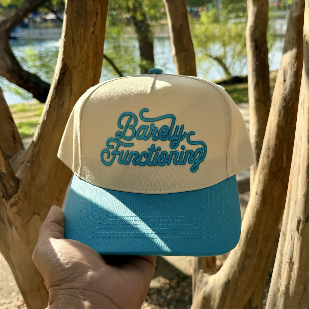 Barely Functioning trucker hat in Natural / Aqua Bill with raised aqua script lettering, held up in front of crape myrtle trees