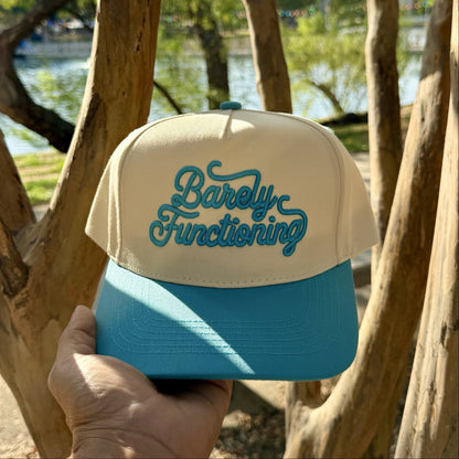 Barely Functioning trucker hat in Natural / Aqua Bill with raised aqua script lettering, held up in front of crape myrtle trees