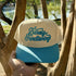 Barely Functioning trucker hat in Natural / Aqua Bill with raised aqua script lettering, held up in front of crape myrtle trees