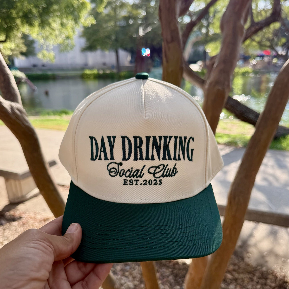 Day Drinking Social Club trucker hat in Natural / Forest Green Bill with forest green lettering, held up by a lake