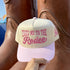 Take Me to the Rodeo trucker hat in Natural / Pink Bill with raised pink lettering, held up in front of giant cowboy boots