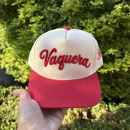 Vaquera trucker hat in Natural / Red Bill with raised red script lettering, held up outdoors in front of green shrubs