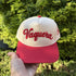 Vaquera trucker hat in Natural / Red Bill with raised red script lettering, held up outdoors in front of green shrubs