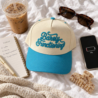 Barely Functioning trucker hat flat lay on a bedsheet with iced coffee, a planner, sunglasses, a phone at 1% battery, and a claw clip