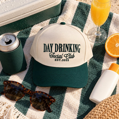 Day Drinking Social Club trucker hat flat lay on a striped towel with sunglasses, a koozie, a mimosa, and an orange slice