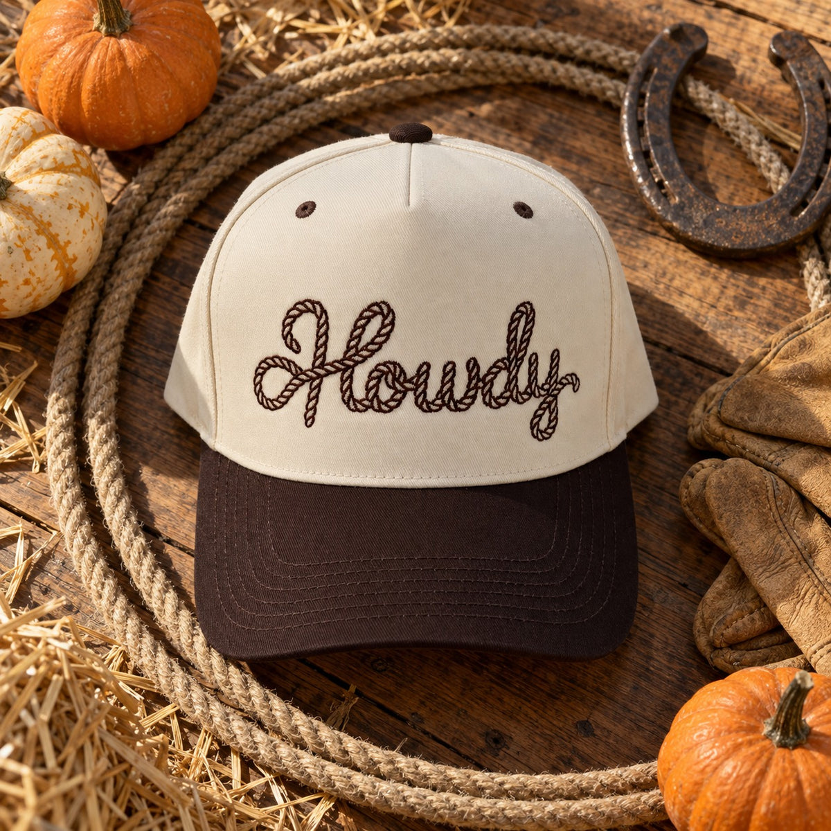 Howdy Rope trucker hat flat lay on a wooden table with a coiled lasso, a horseshoe, leather gloves, pumpkins, and hay