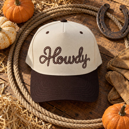 Howdy Rope trucker hat flat lay on a wooden table with a coiled lasso, a horseshoe, leather gloves, pumpkins, and hay