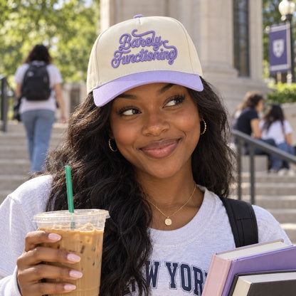 College student with iced coffee wearing the Barely Functioning trucker hat in Natural / Lavender Bill with lavender embroidery