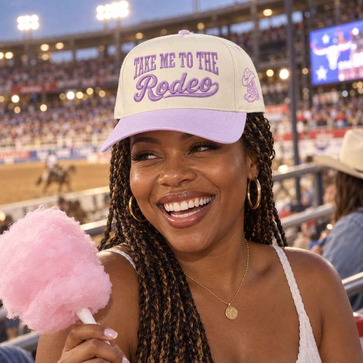 Woman with cotton candy in a rodeo grandstand wearing the Take Me to the Rodeo trucker hat in Natural / Lavender Bill with lavender embroidery
