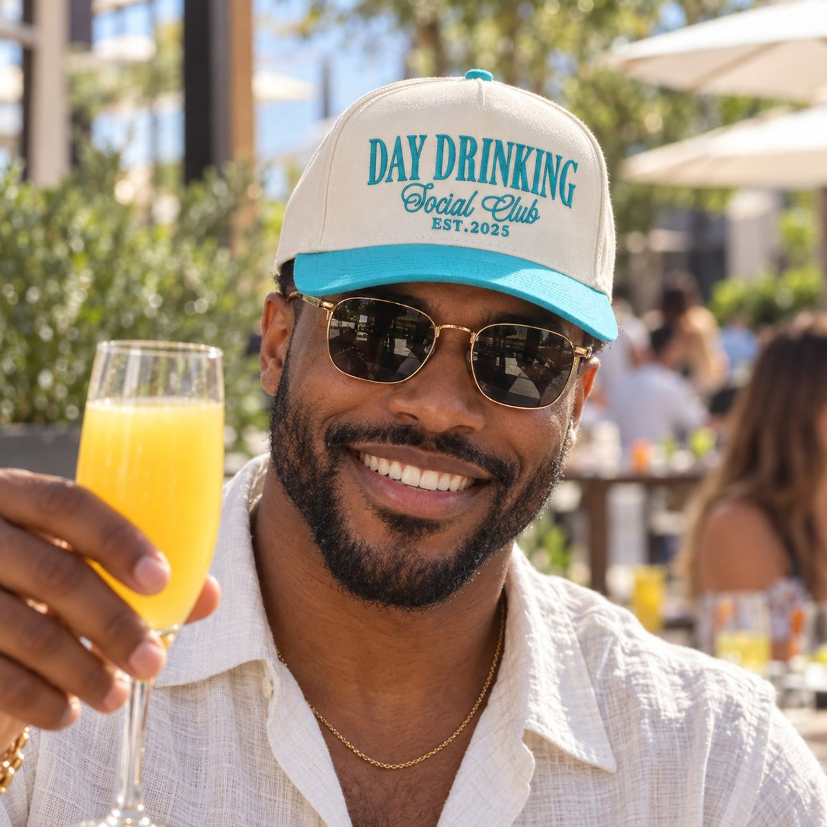 Man with a mimosa on a brunch patio wearing the Day Drinking Social Club trucker hat in Natural / Aqua Bill with aqua embroidery