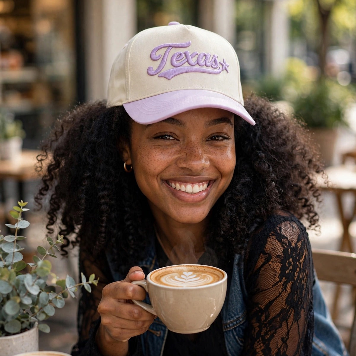 Woman at a sidewalk cafe holding a latte while wearing the Texas 3D Star trucker hat in Natural / Lavender Bill with matching lavender embroidery