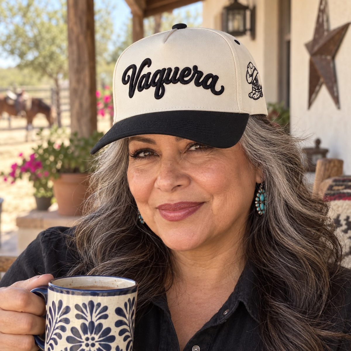 Woman on a ranch porch with a coffee mug wearing the Vaquera trucker hat in Natural / Black Bill with black embroidery