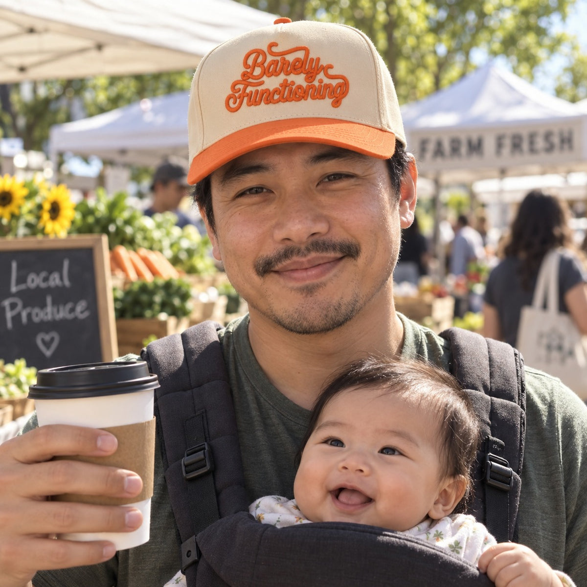 New dad with a baby carrier at a farmers market wearing the Barely Functioning trucker hat in Natural / Orange Bill with orange embroidery