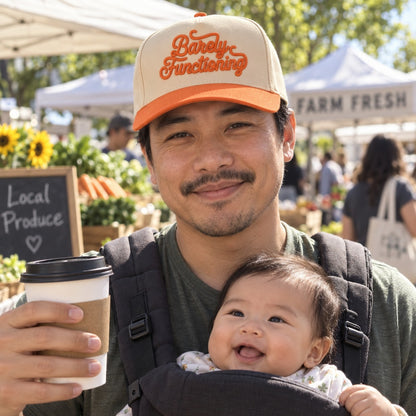 New dad with a baby carrier at a farmers market wearing the Barely Functioning trucker hat in Natural / Orange Bill with orange embroidery