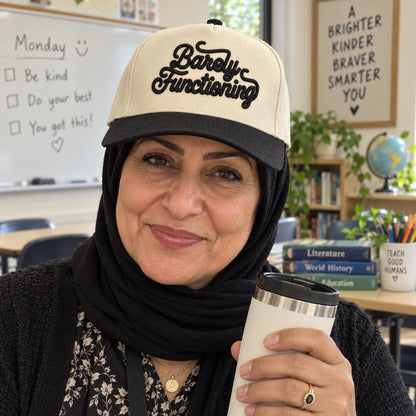 Teacher with a travel mug on a Monday morning wearing the Barely Functioning trucker hat in Natural / Black Bill with black embroidery