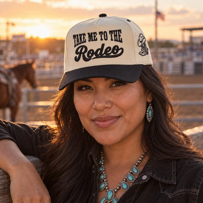 Woman at a rodeo arena fence at sunset wearing the Take Me to the Rodeo trucker hat in Natural / Black Bill with black embroidery