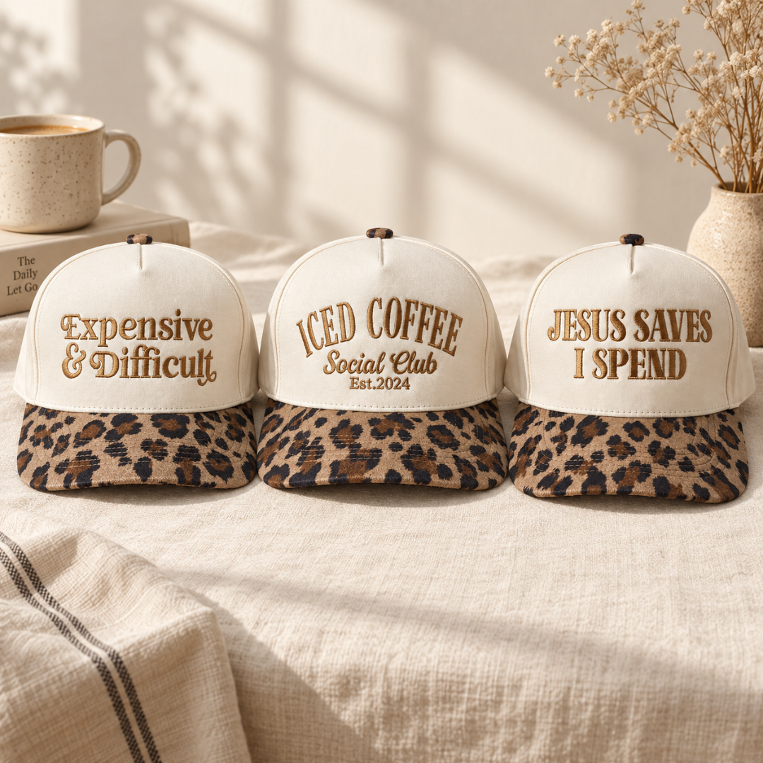 Three beige baseball caps with leopard-print visors are lined up on a beige fabric surface. The left cap reads &