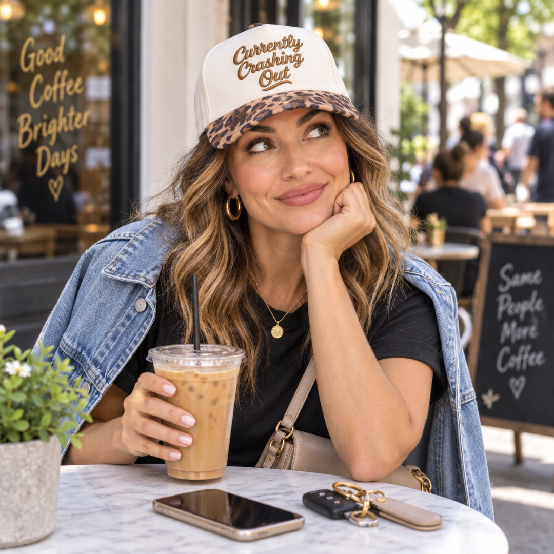 A woman sitting at a cafe table wearing a cream and leopard-print baseball cap with the text &