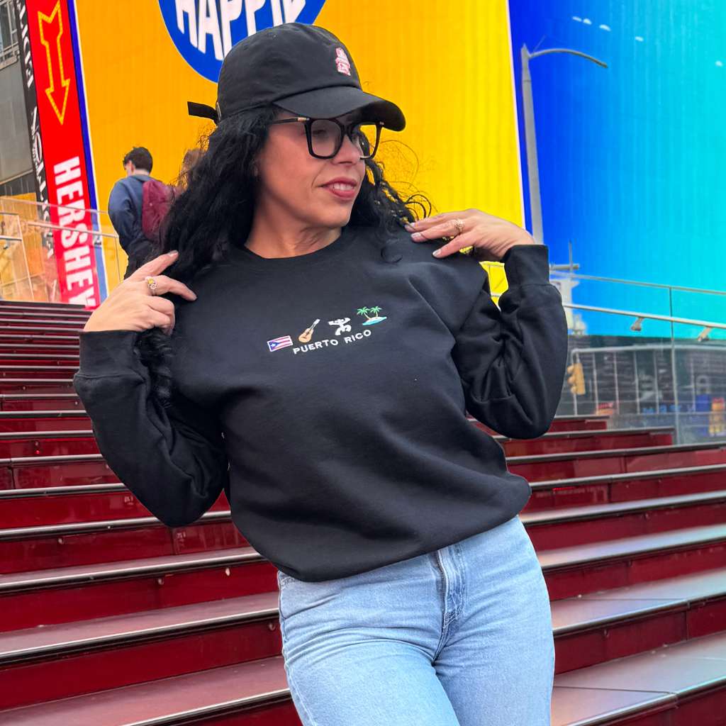Woman wearing the black DTMF Puerto Rico Icons embroidered sweatshirt from DSY Lifestyle, standing on red steps with a colorful background.
