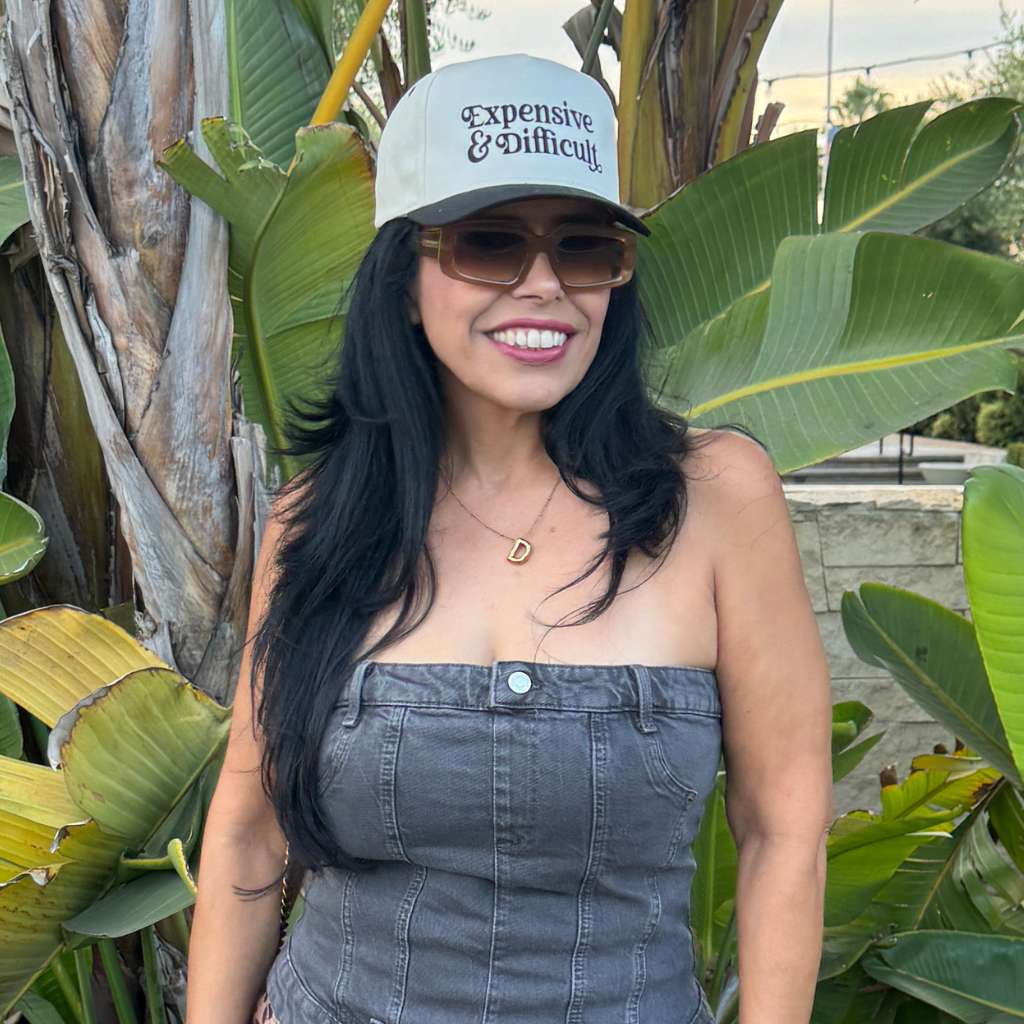 Woman wearing a cap with text, sunglasses, and a strapless denim dress standing among tropical plants embroidered by DSY Lifestyle