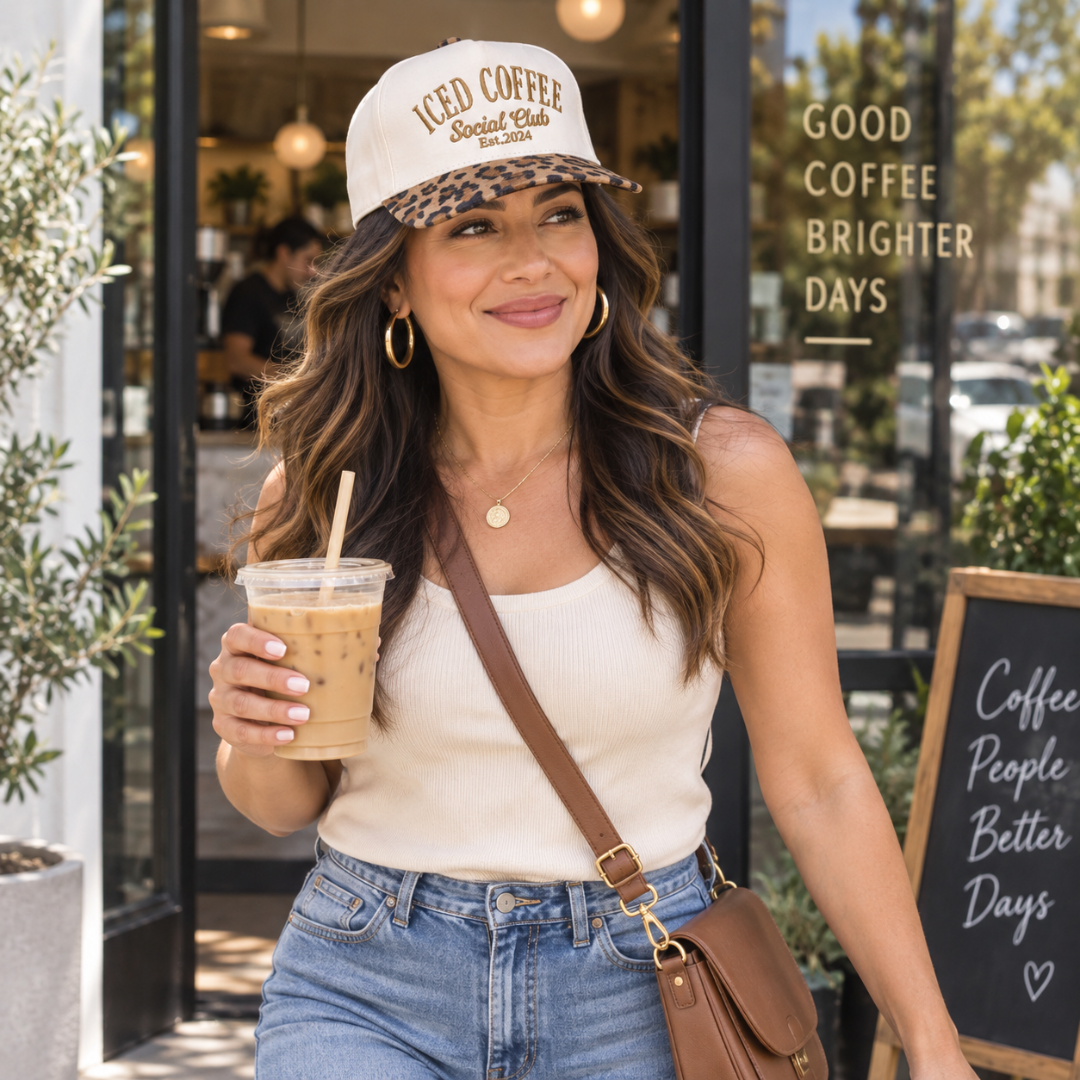 A woman standing outside a coffee shop wearing a cream and leopard-print baseball cap that reads &