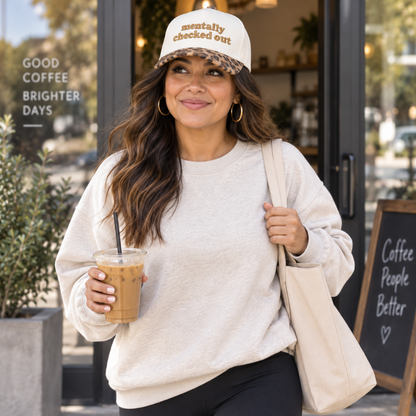 A woman standing outside a coffee shop wearing a cream crewneck sweatshirt, black pants, a beige canvas tote bag, and a white baseball cap with the text &