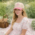Female sitting in park on a blanket wearing a Natural Pink Nail Cross 3D Puff Two-Tone Baseball Hat featuring a raised nail cross on a structured natural crown with a contrasting visor; a premium faith statement by DSY Lifestyle.