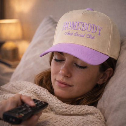 Female relaxing wearing a Natural Lavender Homebody Anti-Social Club embroidered baseball hat. A two-tone statement hat for women who love comfort, style, and effortless outfits. -DSY Lifestyle