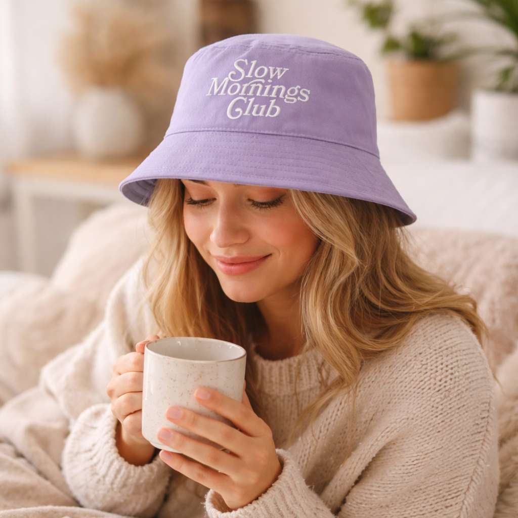 Woman wearing a purple bucket hat with embroidered Slow Mornings Club text, holding a mug indoors.