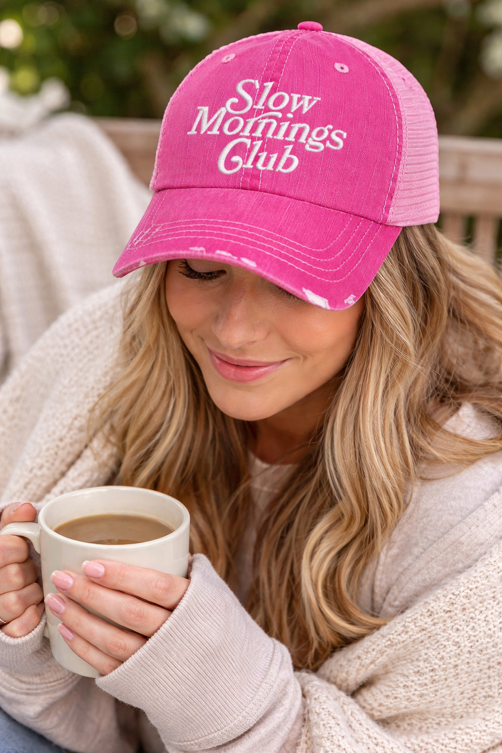 Woman wearing a pink Slow Morning Club'cap and holding a mug, wrapped in a blanket.