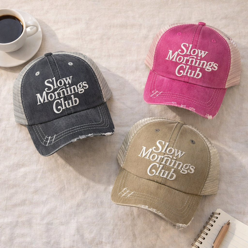Three hats with embroidered with Slow Mornings Club text on a textured surface with a cup of coffee
