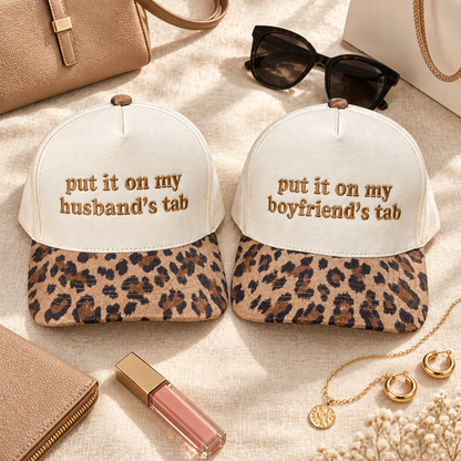 Two matching trucker-style hats displayed on a beige fabric surface with lifestyle accessories. Each hat has an off-white crown with brown embroidered text reading &