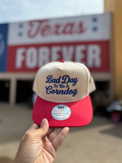 Bad Day To Be A Corndog embroidered hat in Natural with a red bill and royal blue lettering, held up at the fair