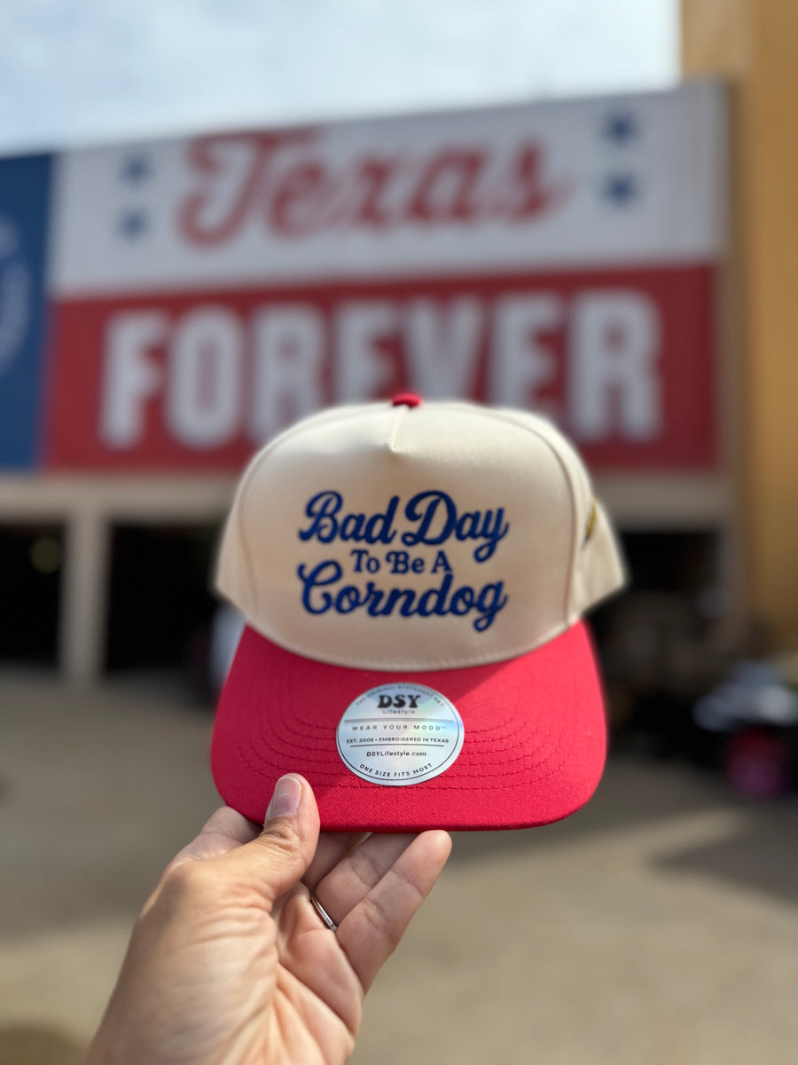 Bad Day To Be A Corndog embroidered hat in Natural with a red bill and royal blue lettering, held up at the fair