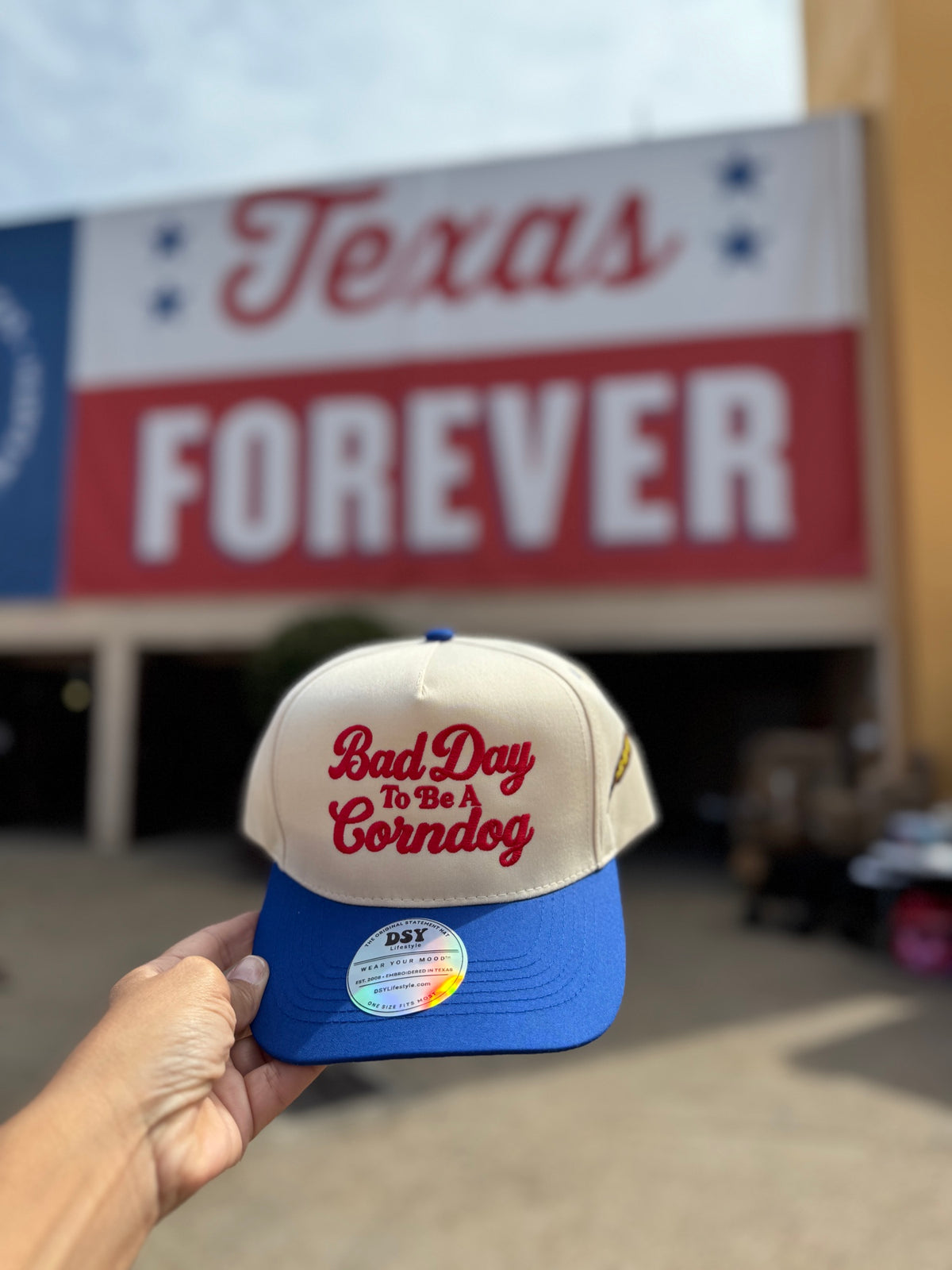 Bad Day To Be A Corndog embroidered hat in Natural with a royal blue bill and red lettering, held up at the fair