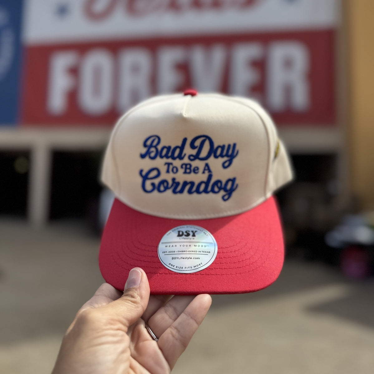 Natural trucker hat with red bill embroidered Bad Day To Be A Corndog, held up in front of the Texas Forever sign at the fair