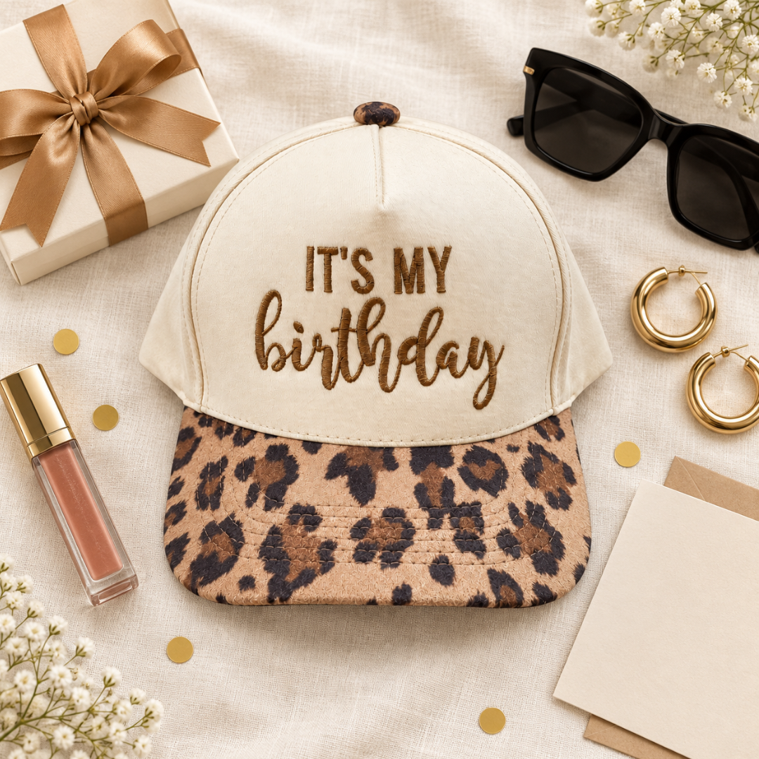 A baseball cap styled in a flat lay with a cream crown and a leopard-print visor. The front of the cap features the embroidered text &