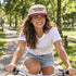 Woman riding a bike in the park wearing the make heaven crowded trucker hat with a natural crown and maroon bill