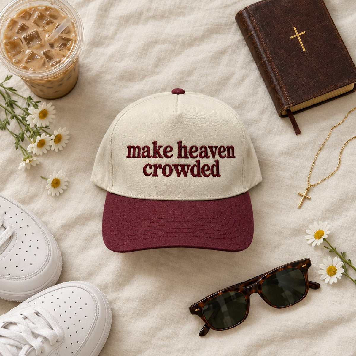 Make heaven crowded trucker hat styled flat lay with a Bible, cross necklace, iced coffee, sneakers, and daisies