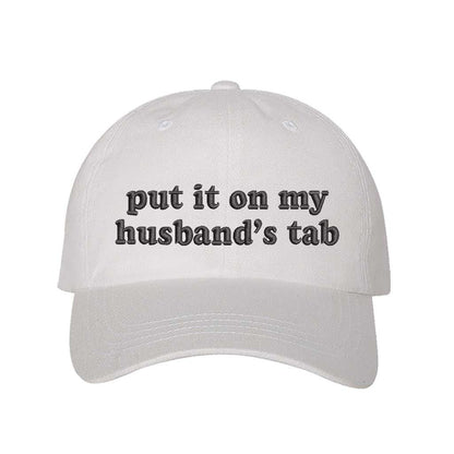 White Put It on My Husband’s Tab Embroidered Baseball Hat in soft cotton with curved brim and adjustable back strap, perfect for vacations and girls’ nights. DSY Lifestyle.