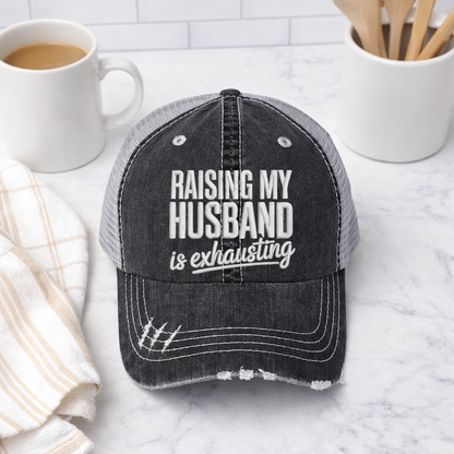 Black flat lay of a DSY Lifestyle distressed trucker hat featuring the phrase Raising My Husband Is Exhausting in bold, professional embroidery. - DSY Lifestyle